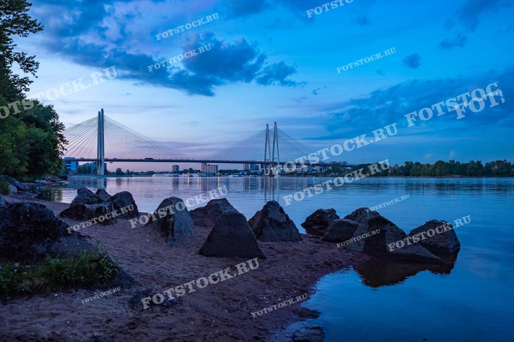 Фотография показывает мост через реку в сумеречное время суток, что создаёт успокаивающую и медитативную атмосферу. Мягкий свет ламп отражается на водной поверхности, а контраст между тёмными камнями на берегу и мягким голубым небом дополняет сцену. Этот снимок может быть использован в архитектурных и туристических публикациях, как часть фотоальбома о Санкт-Петербурге или как элемент декора.